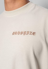 Lyse beige t-shirt med rund hals, prydet med "DROPSIZE" i brun gotisk broderi på forsiden. Glat overflade, afslappet design.