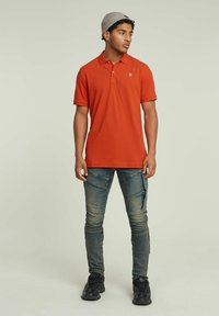 Polo arancione con colletto e patta a tre bottoni, abbinato a jeans blu texturizzati con tasche con cerniera e sneakers nere.