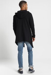 Sudadera negra de manga larga con una gran capucha, que presenta un dobladillo curvado y un corte relajado, combinada con unos vaqueros de un azul claro y zapatillas grises.