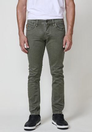Uomo che indossa jeans verde oliva slim fit, t-shirt bianca e sneakers nere, in piedi su uno sfondo chiaro e uniforme.