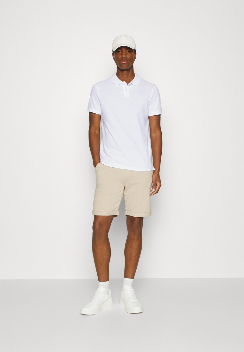 Hvit polo-skjorte, beige shorts og hvite joggesko. Modellen har på seg en hvit caps. Glatt, lett materiale med kortermede skjorter og rullet shorts.
