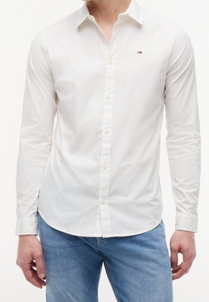 Homme portant une chemise blanche cintrée à manches longues avec un petit logo sur la poitrine et un jean bleu clair.