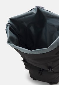 Sac à dos noir avec une fermeture supérieure à enroulement, dotée d'une doublure intérieure grise et de cordons de serrage ajustables. Le matériau semble résistant et texturé.