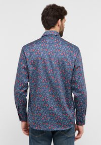 Herenoverhemd van donkerblauwe stof met een bloemenpatroon in het rood, roze en groen. Voorzien van een kraag, lange mouwen en een gebogen zoom.