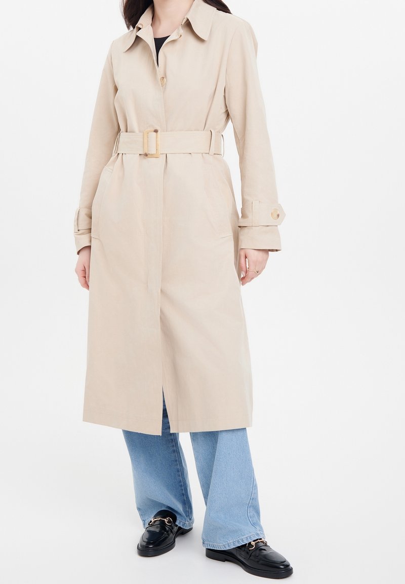 Beige trenchcoat med bälte i midjan, slagkrage, sidofickor och knäppte ärmar. Matchad med utsvängda blå jeans och svarta skor.