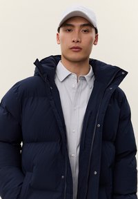 Jauns vīrietis valkā baltu cepuri, gaiši pelēku pogājamu kreklu un tumši zilu polsterētu jaku ar kapuci, raugoties tieši kamerā pret vienkāršu fonu.