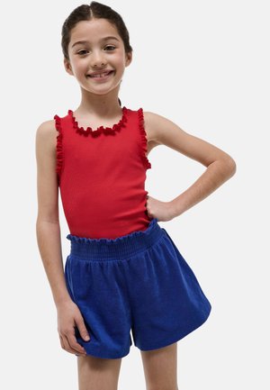 Fille souriante, main sur la hanche, portant un top rouge sans manches avec des volants et un short bleu à taille élastique sur un fond uni.