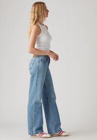 Light blue wide-leg jeans with a high waist, made of denim, featuring a brown leather patch and subtle back pocket stitching. Paired with white tank top.