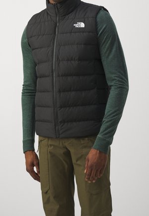 Mężczyzna w czarnej pikowanej kamizelce z logo The North Face, zielonej koszuli z długim rękawem i oliwkowych spodniach typu cargo, stojący na jednolitym tle.