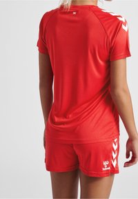 Punainen urheilupaita, jossa on lyhyet hihat, ja siihen sopivat shortsit. Paidassa on valkoiset vinonauha-kuvioinnit hihoissa ja logo alaosassa.