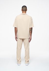 Beige ribbad kortärmad skjorta med krage, tillsammans med matchande ribbade byxor. Slät textur med en avslappnad passform. Vita sneakers.