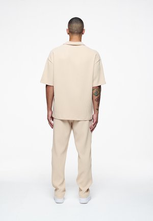 Beige ribbel shirt met korte mouwen en een kraag, gecombineerd met bijpassende ribbelbroek. Soepele textuur met een relaxte pasvorm. Witte sneakers.