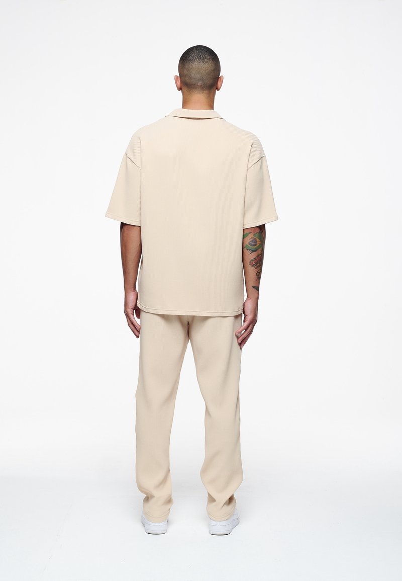 Beige ribbad kortärmad skjorta med krage, tillsammans med matchande ribbade byxor. Slät textur med en avslappnad passform. Vita sneakers.