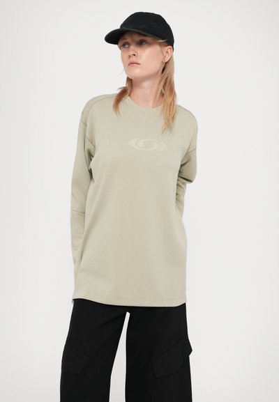 Camisa de manga longa verde-oliva clara com um padrão de malha texturizada, apresentando um logótipo discreto ao centro, combinada com calças cargo pretas e um boné preto.