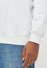 Høyre hånd av en person som har på seg en lysegrå langermet skjorte og blå denimbukser, som hviler avslappet ved siden av seg.