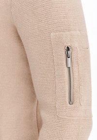 Beżowy sweter z dzianiny z kapturem i kieszenią na zamek błyskawiczny. Tkanina ma teksturowany wzór, a zamek błyskawiczny ma metaliczne wykończenie.