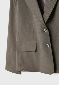 Veste marron et beige à petit motif pied-de-poule, avec deux boutons à l'avant et une poche à rabat sur le côté inférieur.
