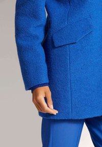 Blauwe wollen jas met een textuurafwerking, voorzien van grote voorzakken en geribbelde manchetten. Het op maat gemaakte ontwerp wordt aangevuld met een bijpassende blauwe outfit.