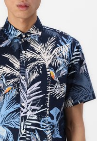 Chemise à manches courtes avec un fond bleu marine orné de motifs de feuilles de palmier vibrantes en bleu et pastel, et d'oiseaux aux accents orange. Fermeture à boutons.