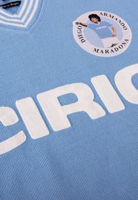 Jersey de punto azul claro con la palabra "CIRIO" en letras blancas prominentes y un parche circular con una ilustración de un jugador y detalles de su nombre.