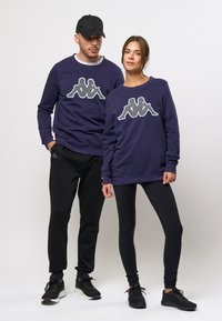 Fialové mikiny s logom Kappa, skombinované s čiernymi nohavicami a teniskami. Mikiny majú dlhé rukávy a okrúhle výstrihy.