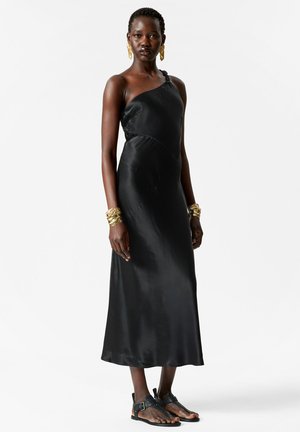 Woman wearing a black one-shoulder satin dress, black sandals, large gold earrings, and chunky gold bracelets, standing on a white background.