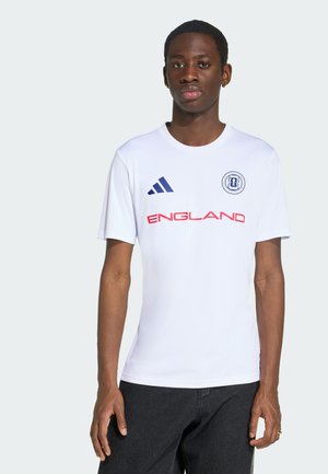 Jeune homme portant un maillot blanc de l'Angleterre avec un logo Adidas bleu et un texte rouge, debout devant un fond clair uni.