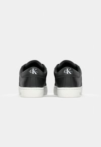 Zapatillas de cuero negro con suelas de goma blanca, que presentan una puntera redonda y un acento de logo metálico en el talón trasero. Textura suave.