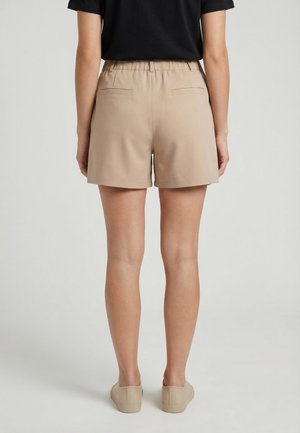 Vista trasera de una persona que lleva pantalones cortos beige a medida, camisa negra metida por dentro y zapatos beige sin cordones contra un fondo liso.