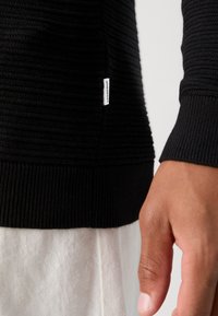 Svart ribbestrikket genser med teksturert stoff. Har en liten hvit etikett ved kanten. Modellen har den på seg over lyse bukser.