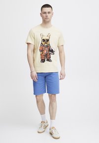 Beige grafisk t-shirt med en fransk bulldog i en orange astronautdragt, parret med blå shorts og hvide sneakers.
