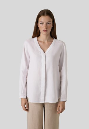 Frau mit glatten braunen Haaren, die eine weiße langärmelige V-Ausschnitt-Bluse und beige Hose trägt, steht vor einem schlichten hellgrauen Hintergrund.