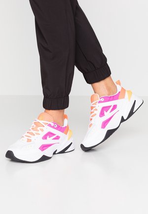 Personne portant des baskets blanches avec des accents roses, orange et jaunes et un pantalon noir retroussé debout sur un sol lisse gris clair.