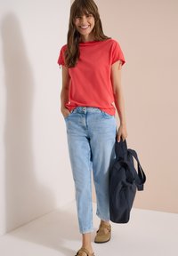 Femme souriante en haut rouge à manches courtes et jean bleu clair, tenant un sac cabas bleu marine et se tenant devant un fond beige.