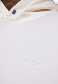 Hoodie blanc à côtes avec poche frontale, capuche à cordon et logo embossé "ESTEEM" sur la poitrine dans une couleur assortie.