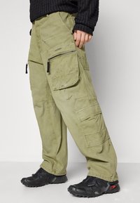 Person wearing olive green cargo pants with multiple pockets and black athletic shoes, standing against a plain light background.