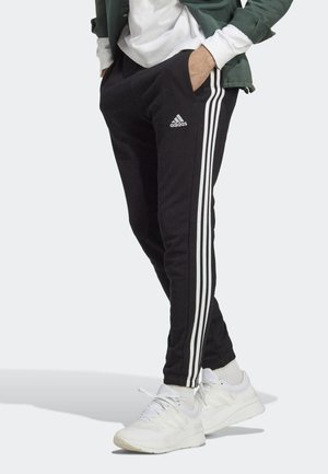 Persoon draagt zwarte Adidas trainingsbroek met witte strepen en logo, witte sneakers, groene jas en wit sweatshirt met handen in zakken.