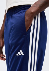 Άτομο που φοράει μπλε φόρμα Adidas με λευκές ρίγες και λογότυπο, το χέρι ακουμπά στην εμπρόσθια τσέπη.