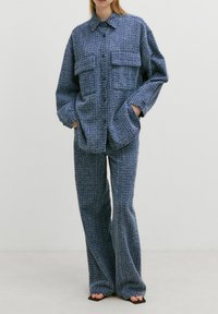 Blauwe textuur shirtjasje met twee opgestikte zakken aan de voorkant, gecombineerd met bijpassende wijde broek. Het materiaal lijkt geweven, zacht en lichtgewicht.