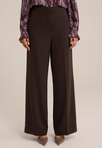 Woman wearing high-waisted, wide-leg dark brown pants, paired with taupe pointed-toe heels and a long-sleeve patterned blouse.