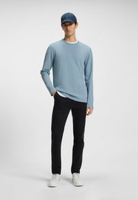 Light blue long-sleeve shirt with round neckline and textured fabric paired with black trousers and white sneakers, and a dark blue cap.