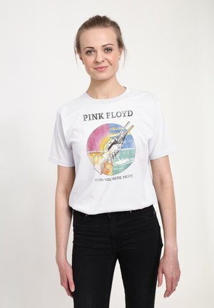 Femme portant un t-shirt blanc Pink Floyd « Wish You Were Here » avec un design coloré de poignée de main, associé à un pantalon noir, sur un fond uni.