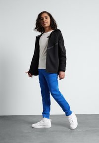 Black zip-up hoodie with gray accents over a white t-shirt, paired with blue sweatpants and white sneakers. Smooth fabric and casual fit.