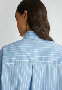 Camisa de rayas azul claro con líneas verticales blancas, con un cuello alto, pliegue trasero y un pequeño acento metálico cerca del cuello.