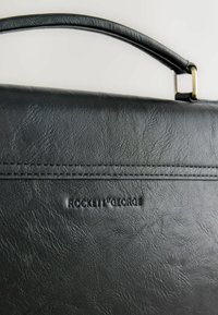 Czarna torba z imitacji skóry o gładkiej fakturze, wyposażona w górny uchwyt i tłoczone logo. Proste przeszycia wzdłuż szwów dodają subtelnego detalu.