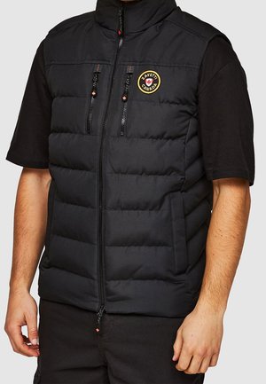 Homme portant un gilet sans manches matelassé noir avec deux poches poitrine zippées verticales et un écusson circulaire portant l'inscription "ZAVETTI CANADA."