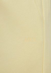 Una tela amarilla claro con una textura suave presenta un sutil logotipo en relieve "JUXX." El diseño incluye un bolsillo lateral.