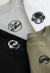 Vier T-Shirts: grau mit dem "Skate of Mind"-Logo, schwarz mit einem Totenkopf und Kopfhörern, weiß mit einem Gorilla, der Kopfhörer trägt, und olivgrün mit einem Gamecontroller.