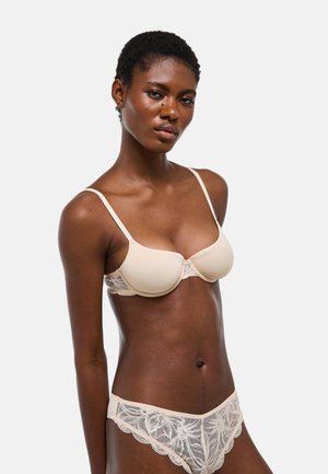 Femme aux cheveux courts portant un soutien-gorge beige à fines bretelles et une culotte en dentelle assortie sur un fond clair uni.