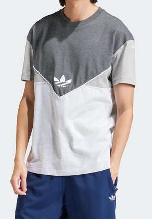 Personne portant un t-shirt Adidas gris et blanc à blocs de couleurs avec un logo sur la poitrine et un pantalon Adidas bleu marine avec un logo sur la poche.